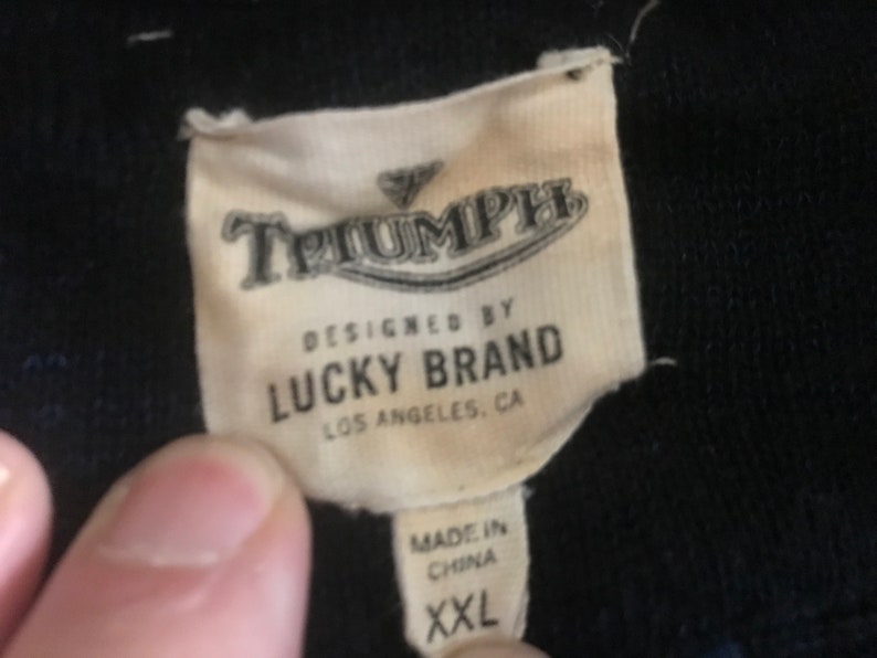 Op de afbeelding: Een wit label met zwarte tekst dat "Triumph Designed by Lucky Brand Los Angeles, CA Made in China XXL" zegt.