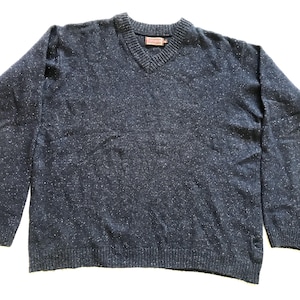 Puede incluir: Un suéter azul marino con cuello en V y un patrón moteado. El suéter tiene cuello, puños y dobladillo acanalados. Una pequeña etiqueta rectangular es visible cerca del escote. El suéter está hecho de un material suave y texturizado.