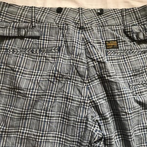 May include: Close-up of the back of plaid pants in shades of gray, black, and blue. The pants have belt loops with buckles, two back pockets, and a small tag with text. The fabric has a woven texture.