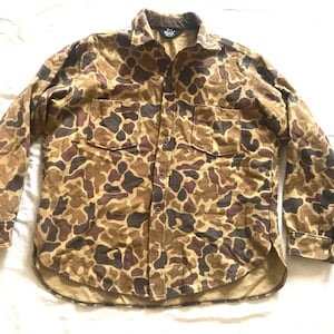 May include: A button-down shirt with a camouflage pattern in shades of brown, tan, and dark brown. The shirt has two chest pockets and a collar. The fabric appears to be a soft, brushed material. The shirt is laid flat on a white surface.