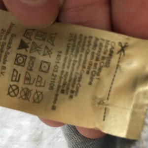 May include: A beige clothing care label with instructions for washing, drying, and ironing. The label also includes the brand name "Scotch & Soda" and the company's address in the Netherlands. The label is made in China.