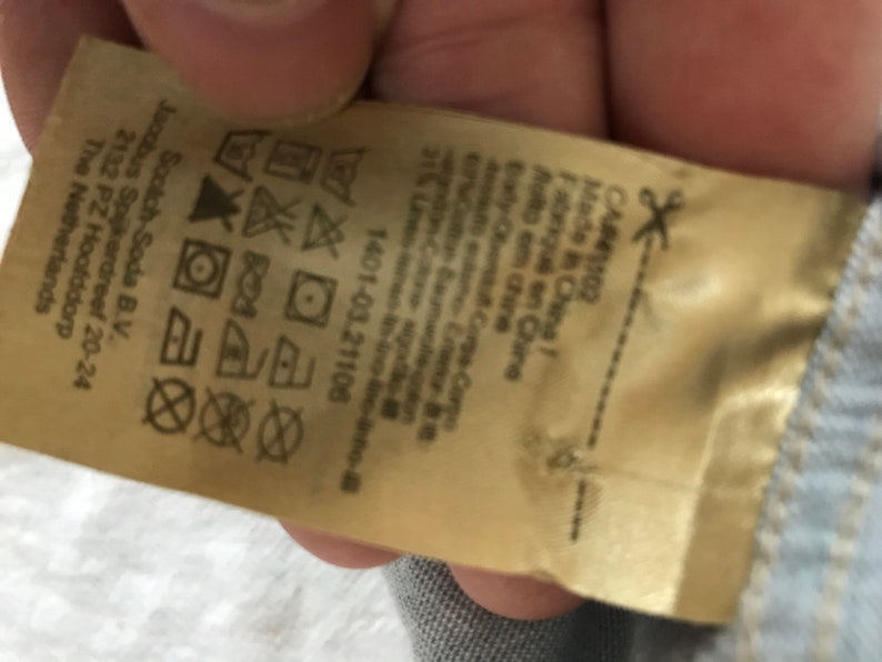May include: A beige clothing care label with instructions for washing, drying, and ironing. The label also includes the brand name "Scotch & Soda" and the company's address in the Netherlands. The label is made in China.