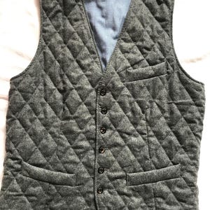May include: A gray quilted vest with a V-neck and button closure. The vest has two front pockets and a light blue lining. The diamond-patterned stitching adds texture to the garment. A classic outerwear piece.