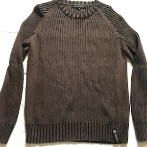 Puede incluir: Un suéter de punto acanalado marrón con cuello redondo y mangas largas. El suéter tiene un patrón de canalé vertical texturizado y una pequeña etiqueta con el logotipo cerca del dobladillo inferior. La prenda es de material suave.