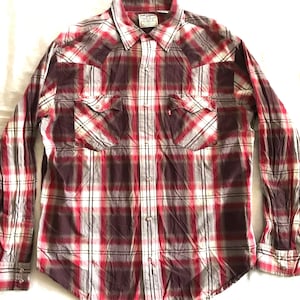 Peut inclure: Une chemise à carreaux à manches longues dans les tons de rouge, blanc et bordeaux. La chemise comporte deux poches de poitrine, une fermeture boutonnée sur le devant et un col. L'étiquette Levi Strauss & Co. est visible en haut.