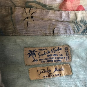 May include: Close-up of two fabric labels sewn into a light blue shirt. The top label is beige with a palm tree graphic and reads "ORIGINAL Scotch & Soda AMS.MFG. CO. Amsterdam Couture First Quality Clothing". The bottom label is beige and reads "Tailor Made Inc. SCOTCH & SODA".
