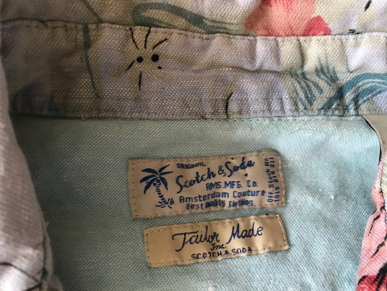 May include: Close-up of two fabric labels sewn into a light blue shirt. The top label is beige with a palm tree graphic and reads "ORIGINAL Scotch & Soda AMS.MFG. CO. Amsterdam Couture First Quality Clothing". The bottom label is beige and reads "Tailor Made Inc. SCOTCH & SODA".