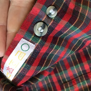 Puede incluir: Primer plano del pu&ntilde;o de una camisa a cuadros rojos, verdes y negros con dos botones grises. Una etiqueta blanca con un c&iacute;rculo verde y s&iacute;mbolos de lavado est&aacute; adjunta. La tela de la camisa es visible.