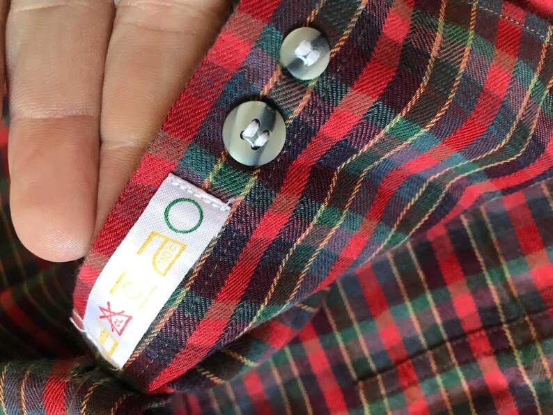Puede incluir: Primer plano del pu&ntilde;o de una camisa a cuadros rojos, verdes y negros con dos botones grises. Una etiqueta blanca con un c&iacute;rculo verde y s&iacute;mbolos de lavado est&aacute; adjunta. La tela de la camisa es visible.
