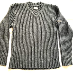 Puede incluir: Un suéter de punto acanalado gris y negro con cuello en V. El suéter de manga larga presenta un patrón de rayas verticales y un pequeño detalle metálico en la manga izquierda. El suéter está hecho de un material grueso y cálido.