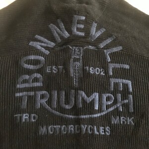 Op de afbeelding: Zwarte gebreide trui met een blauw geborduurd Triumph Motorcycles logo. Het logo bevat de woorden "Bonneville" en "Triumph" in een cirkelvormig ontwerp, met het jaar "1902" en de woorden "TRD" en "MRK" eronder.