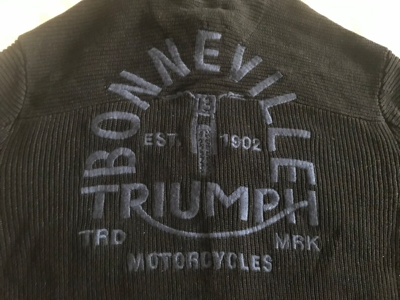 Op de afbeelding: Zwarte gebreide trui met een blauw geborduurd Triumph Motorcycles logo. Het logo bevat de woorden "Bonneville" en "Triumph" in een cirkelvormig ontwerp, met het jaar "1902" en de woorden "TRD" en "MRK" eronder.