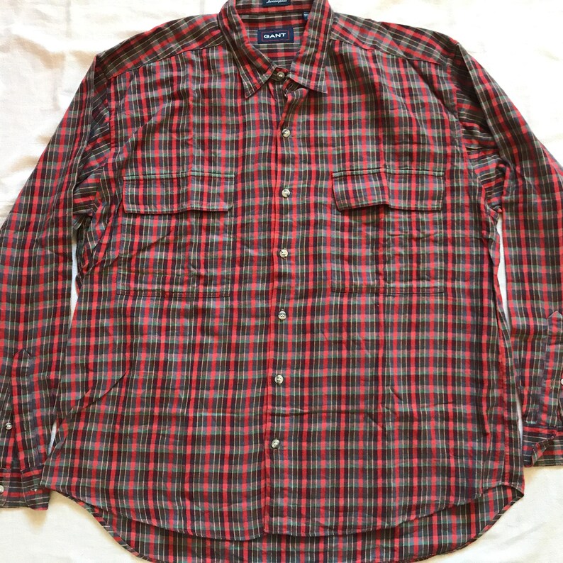Puede incluir: Camisa de manga larga a cuadros rojos y negros. Tiene dos bolsillos en el pecho y la marca "GANT" visible en el cuello. Es una camisa abotonada hecha de tela ligera.