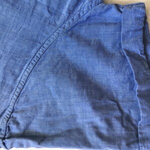May include: Close-up of a blue denim shirt. The fabric has a textured appearance with visible stitching along the seams. The cuff is folded up, revealing the inside of the fabric. The overall look is casual and comfortable.