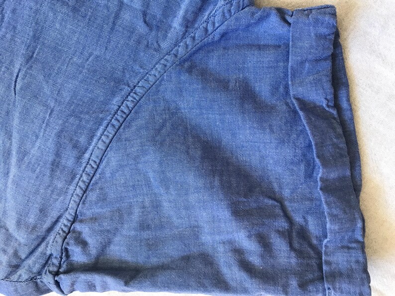 May include: Close-up of a blue denim shirt. The fabric has a textured appearance with visible stitching along the seams. The cuff is folded up, revealing the inside of the fabric. The overall look is casual and comfortable.