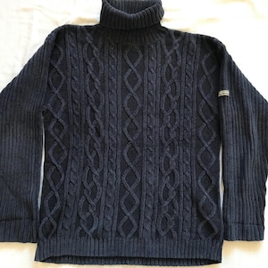 Puede incluir: Un suéter azul oscuro de cuello alto con un patrón de punto de cable. El suéter tiene una textura acanalada y un cuello alto.