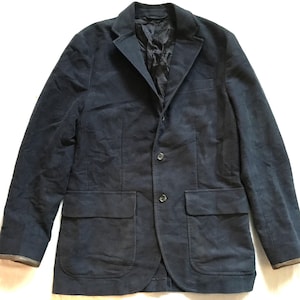 Puede incluir: Una chaqueta azul oscuro con cierre de botones y dos bolsillos de solapa. La chaqueta tiene una tela texturizada y un aspecto ligeramente desgastado.