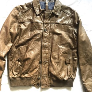 May include: Brown leather jacket with a button-up front, two front pockets, and a ribbed hem and cuffs. The jacket has a collared neckline and a dark blue lining. A classic outerwear piece.