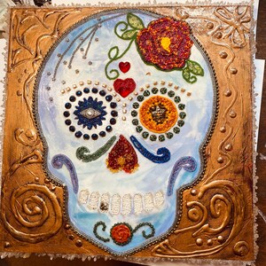 May include: A colorful sugar skull with a blue and white eye, red and gold hearts, and a yellow and green flower. The skull is surrounded by a gold frame with intricate designs.