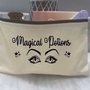 May include: A white canvas makeup bag with a black zipper and the text "Magical Potions" printed in black cursive font. Two black stylized eyes with eyelashes and stars are printed on the bag.