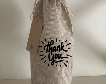 May include: A beige burlap wine bottle bag with a brown paper tag and black lettering that says "Thank You".