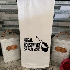 May include: White kitchen towel with black text that reads "The Real Housewives of East York" with a black illustration of a pot and a ladle.
