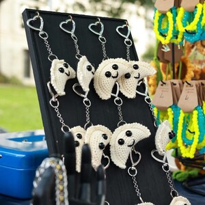 May include: A display of handmade crochet dumpling keychains with smiling faces and heart-shaped clasps. The dumplings are off-white with black eyes and are attached to silver chains. Other colorful beaded bracelets are in the background.