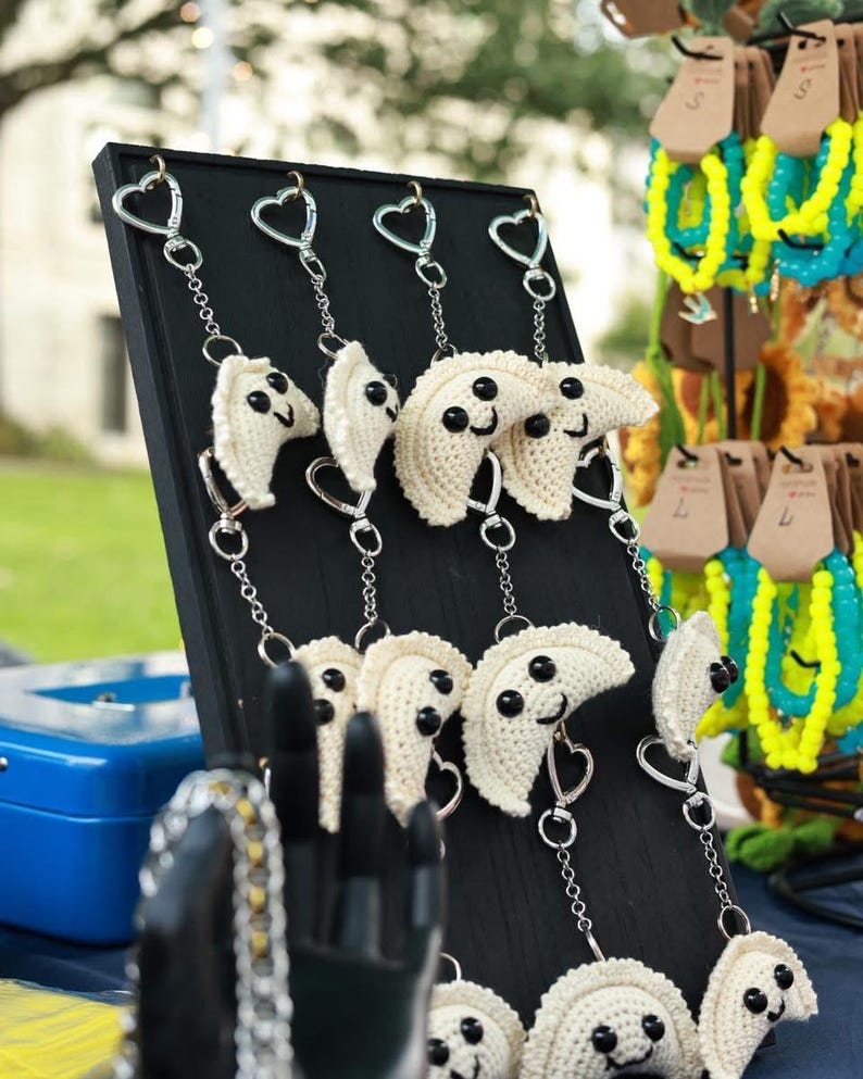 May include: A display of handmade crochet dumpling keychains with smiling faces and heart-shaped clasps. The dumplings are off-white with black eyes and are attached to silver chains. Other colorful beaded bracelets are in the background.