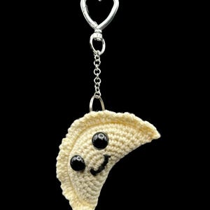 May include: A crocheted dumpling keychain with a smiling face and black bead eyes. The keychain has a silver heart-shaped clasp and chain, perfect for attaching to keys or bags. The dumpling is off-white.
