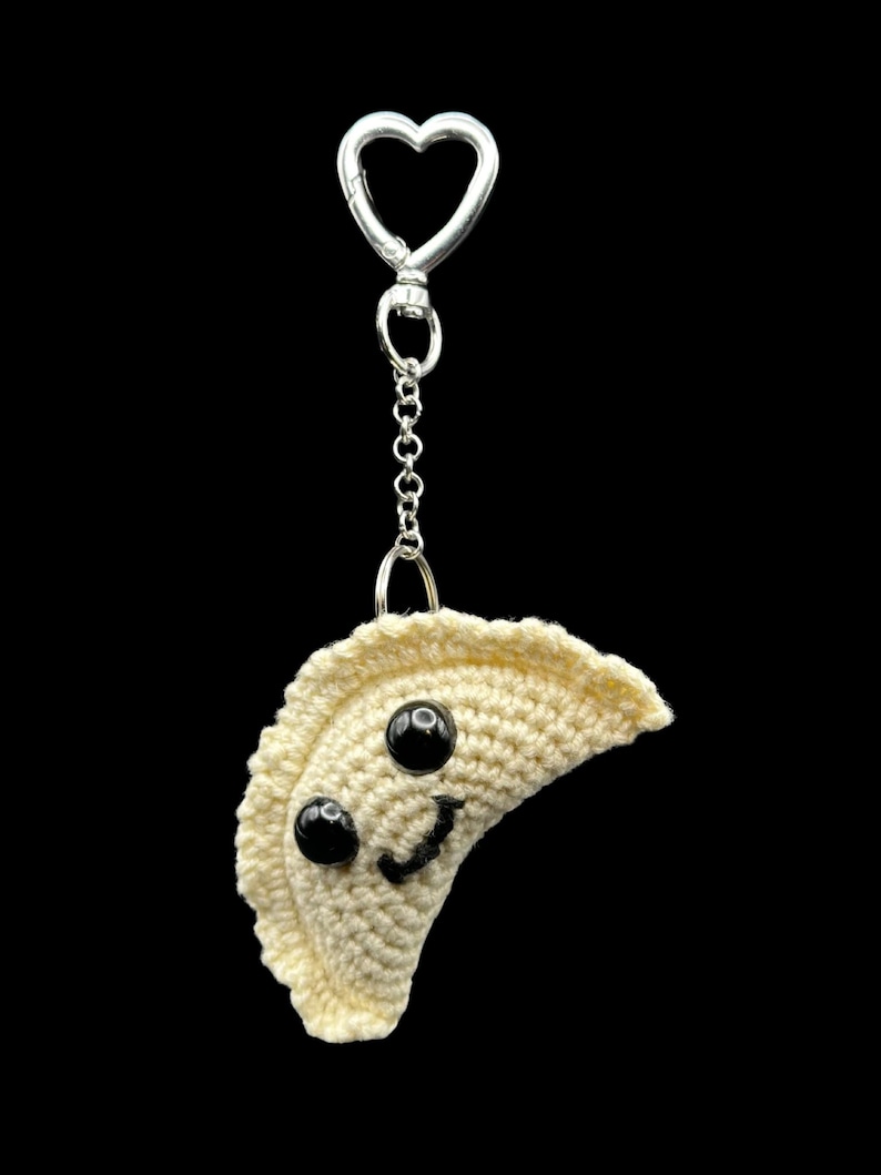 May include: A crocheted dumpling keychain with a smiling face and black bead eyes. The keychain has a silver heart-shaped clasp and chain, perfect for attaching to keys or bags. The dumpling is off-white.