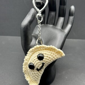 May include: A crocheted dumpling keychain with a smiling face made of black beads and a stitched mouth. The keychain has a silver heart-shaped clasp and chain.