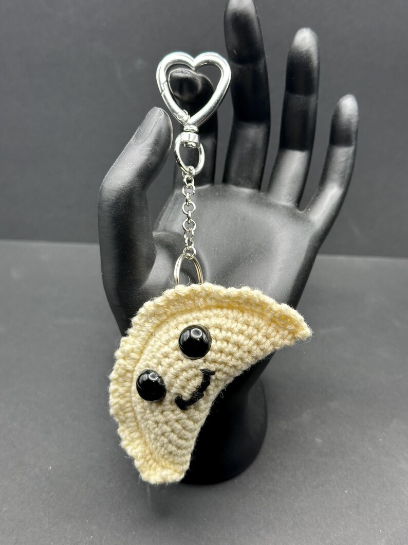 May include: A crocheted dumpling keychain with a smiling face made of black beads and a stitched mouth. The keychain has a silver heart-shaped clasp and chain.