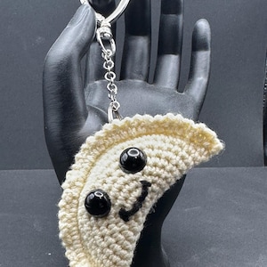 May include: A crocheted keychain in the shape of a dumpling, featuring black bead eyes and a stitched smile. The dumpling is off-white, attached to a silver-colored heart-shaped clasp and chain.