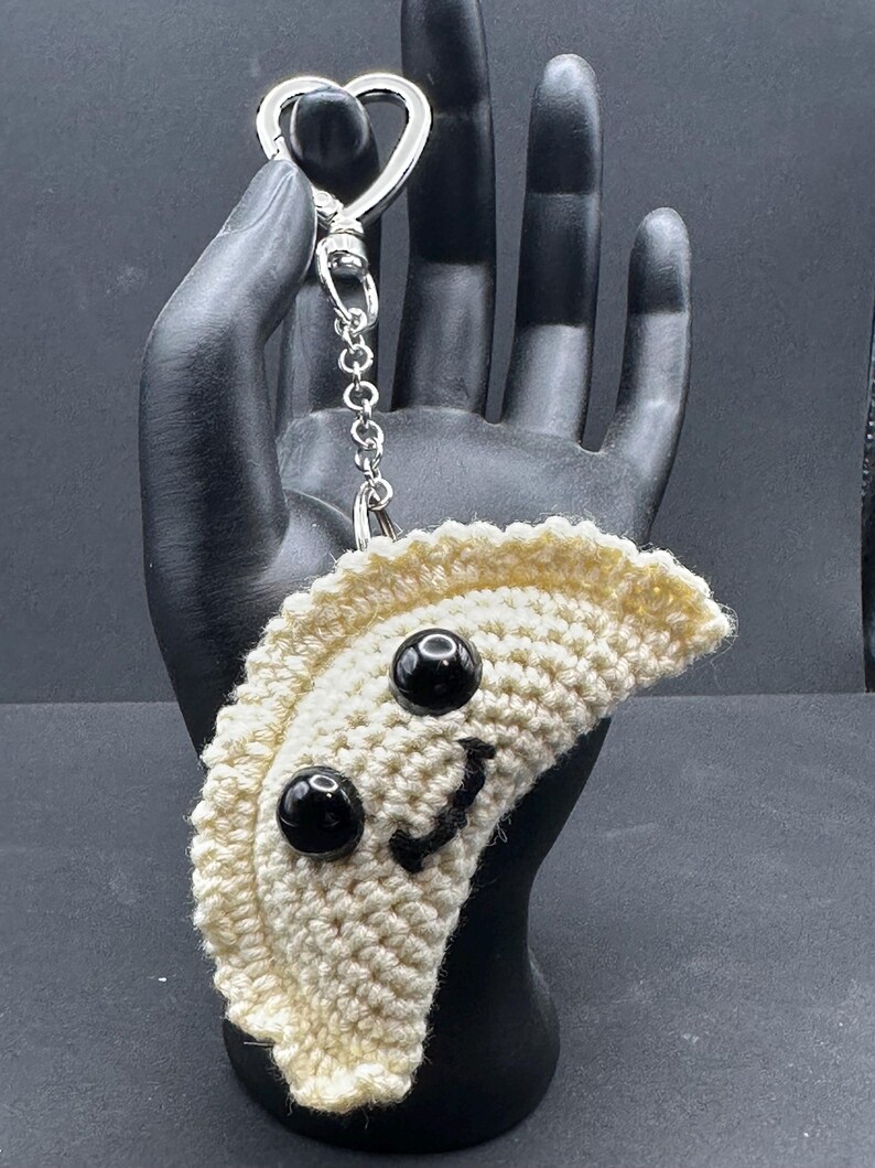 May include: A crocheted keychain in the shape of a dumpling, featuring black bead eyes and a stitched smile. The dumpling is off-white, attached to a silver-colored heart-shaped clasp and chain.