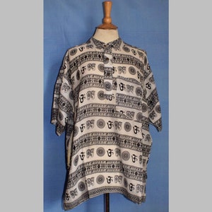Puede incluir: Una camisa blanca de manga corta con un patrón blanco y negro de símbolos Om y otros diseños. La camisa tiene un cuello abotonado y un solo botón de cierre.