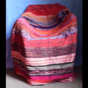 May include: A colourful striped woven throw blanket in shades of red, pink, purple, orange, and brown. The blanket is folded and draped over a chair.