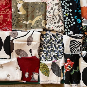 May include: A collection of folded fabric pieces with various patterns. Designs include foxes, leaves, berries, and abstract shapes in colors like red, black, white, and green. Some fabrics have text, and the overall aesthetic is Scandinavian-inspired.