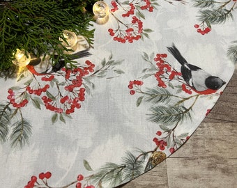 May include: A white round tablecloth with a wintery design featuring red berries, green pine branches, and black and white birds. The tablecloth has a scalloped edge.