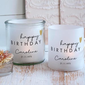 Könnte beinhalten: Zwei weiße Kerzen mit dem Text "happy BIRTHDAY Caroline 21.11.1975" in schwarzer Schrift. Ein kleines goldenes Herz befindet sich neben dem Wort "birthday". Eine Kerze befindet sich in einem klaren Glas, die andere ist ein mattierter Glas-Teelichthalter.