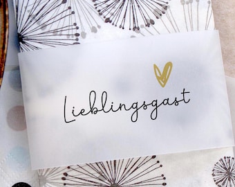 May include: A white card with the word "Lieblingsgast" in black script and a gold heart. The card is on a patterned surface with black and gray dandelion-like designs.
