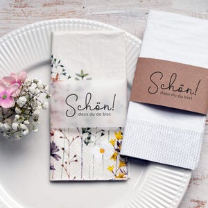 Könnte beinhalten: Zwei weiße Servietten mit einem braunen Papierband. Das Band trägt den Text "Schön! dass du da bist". Eine Serviette hat ein Blumenmuster und ein weißes Etikett mit dem gleichen Text.