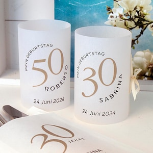 Könnte beinhalten: Zwei weiße Papierzylinder-Kerzenhalter mit einem goldenen Kreisdesign. Der Text auf dem linken Kerzenhalter lautet "Mein Geburtstag 50 Roberto 24. Juni 2024". Der Text auf dem rechten Kerzenhalter lautet "Mein Geburtstag 30 Sabrina 24. Juni 2024".