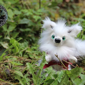 Peut inclure: Un animal en peluche blanc et moelleux avec des yeux verts et un nez noir. L'animal porte un tissu rouge et blanc à motifs autour de sa taille. L'animal est assis dans un lit d'herbe verte.