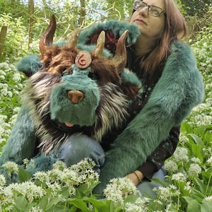 Puede incluir: Una criatura de peluche detallada con pelaje turquesa y marrón, cuernos grandes y una melena esponjosa. La criatura se sostiene en un campo de flores blancas y follaje verde. Un emblema circular es visible en la frente.