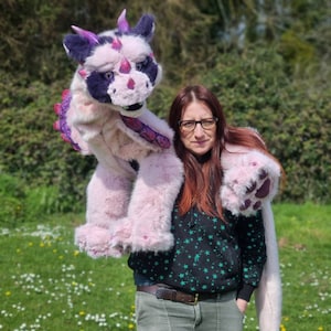 Puede incluir: Una mujer sostiene una gran marioneta de dragón de peluche rosa y morado. El dragón tiene un pelaje rosa y morado con una nariz y ojos negros. La mujer lleva una camiseta negra con estrellas y un par de vaqueros.