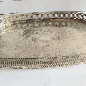 Peut inclure: Un plateau de service ovale en métal argenté présentant un motif floral gravé complexe sur la base, un rebord galerie surélevé avec un design en treillis ajouré vertical, deux poignées latérales et quatre petits pieds décoratifs.