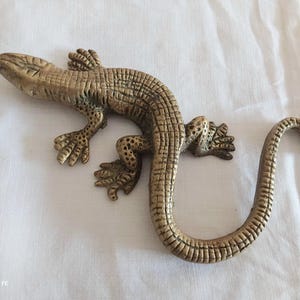 May include: A brass lizard figurine featuring a textured, scaled surface across its body, head, and long curved tail. The metal reptile is depicted in a crawling position with four distinct feet, each showing detailed toes and a weathered finish.