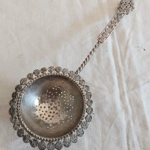 May include: A silver-toned metal tea strainer featuring a circular bowl with a perforated base and a decorative scalloped edge. The handle is long and slender with a twisted metal section and a flat, rectangular end piece with embossed details.