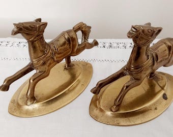 May include: Two gold-toned metal horse figurines on oval bases. The horses are in a running pose with their heads turned to the side.