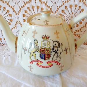 Op de afbeelding: Een witte theepot van keramiek met een gouden rand en een koninklijk wapen met een leeuw en een eenhoorn. Het wapen bevat de tekst "E II R" en "Coronation of Queen Elizabeth II June 2nd 1953".